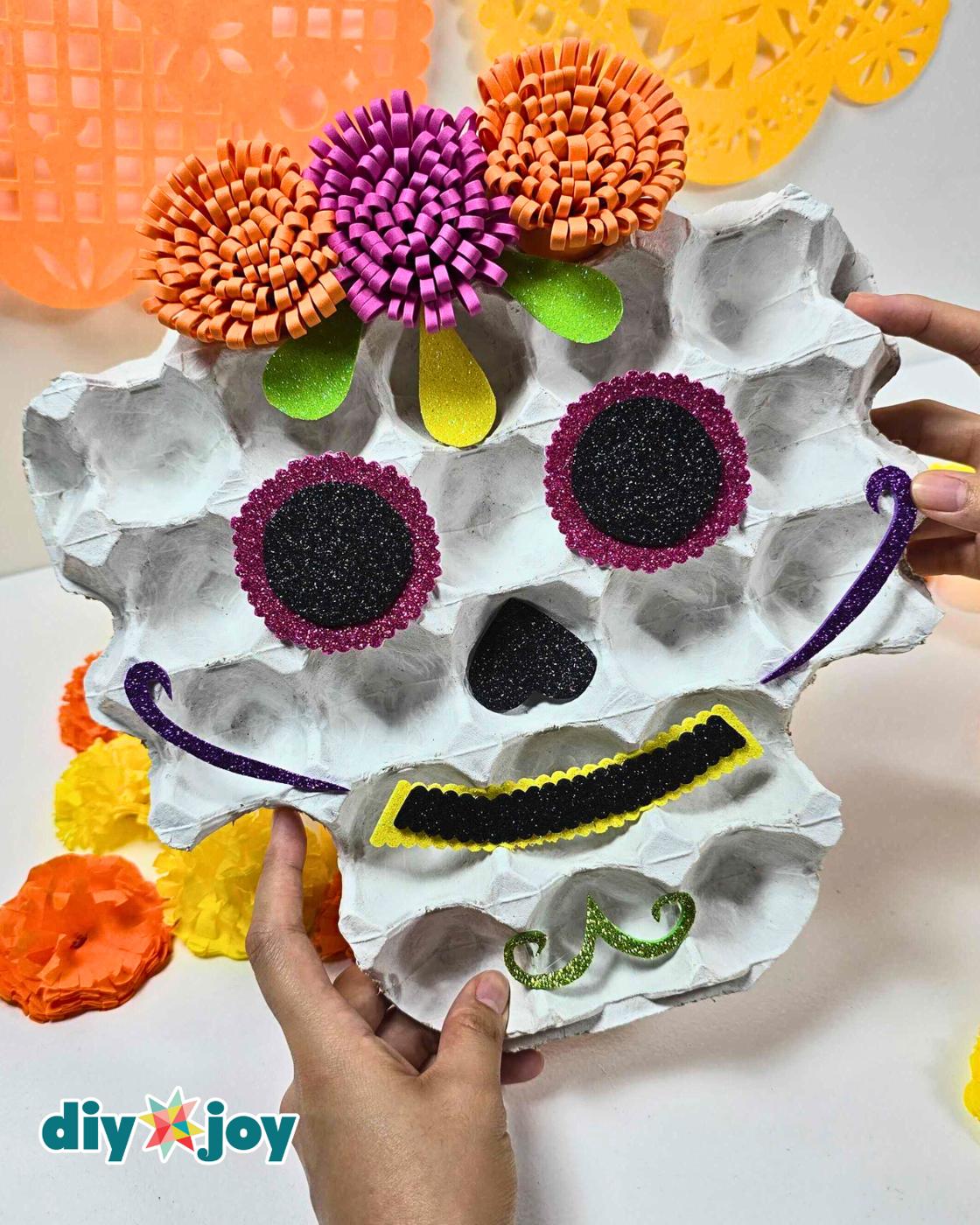 Easy kids crafts - two hands holding the finished glitter foam sugar skull close to the camera in front of orange and purple papel picado