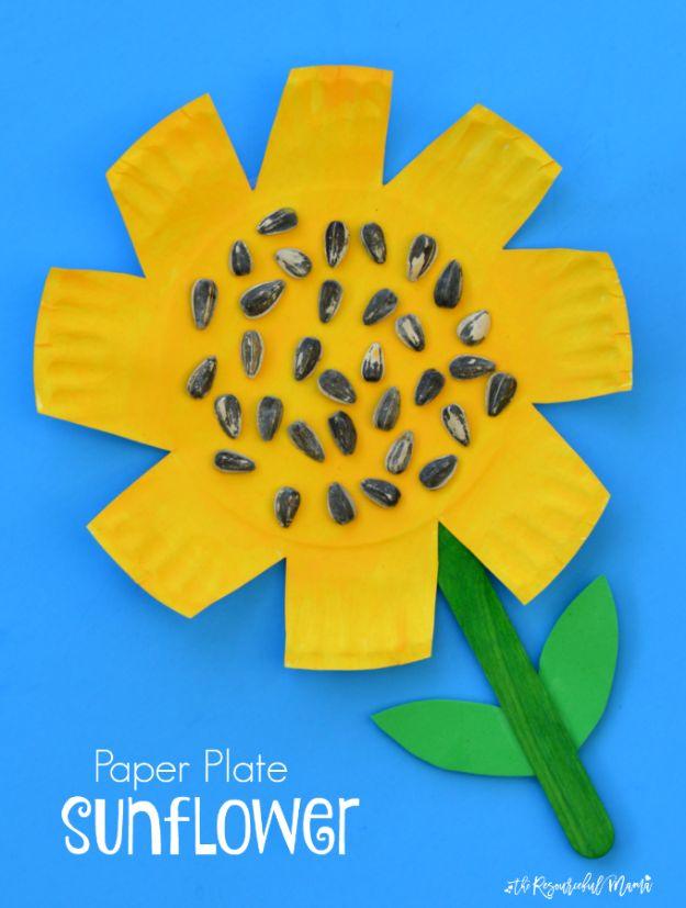 Yellow painted paper plate sunflower with real seeds in the center, a green craft stick stem and foam leaves on blue