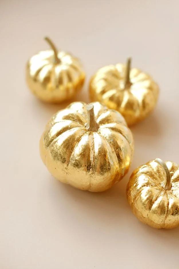 Four small pumpkins covered entirely in shiny gold leaf, grouped together on a plain pale gray background