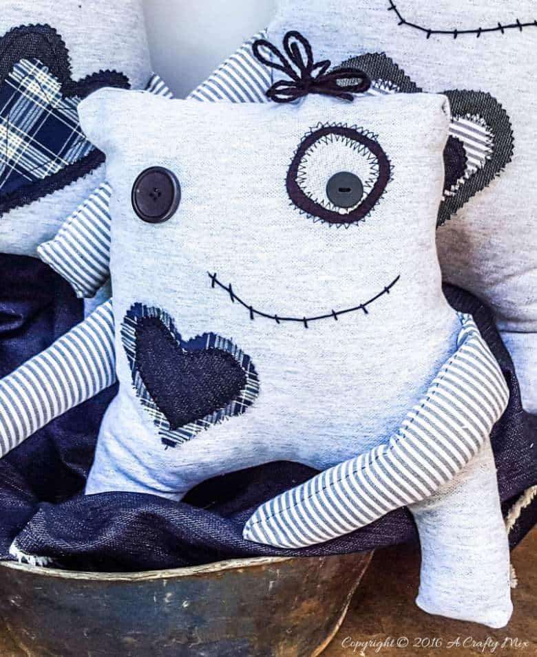 A gray fabric monster softie with one button eye, a stitched smile, a denim heart and striped floppy arms in a metal bowl