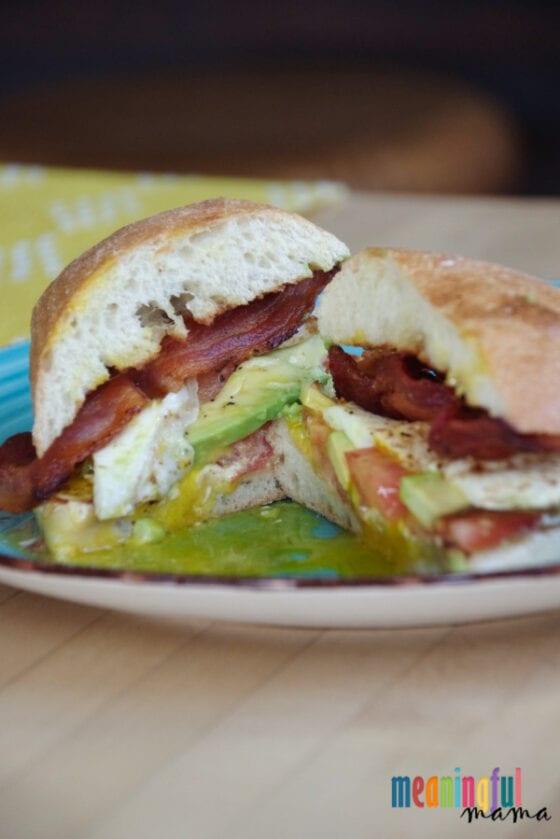 Breakfast sandwich on a ciabatta roll with bacon, avocado, tomato and a fried egg on a green and yellow plate