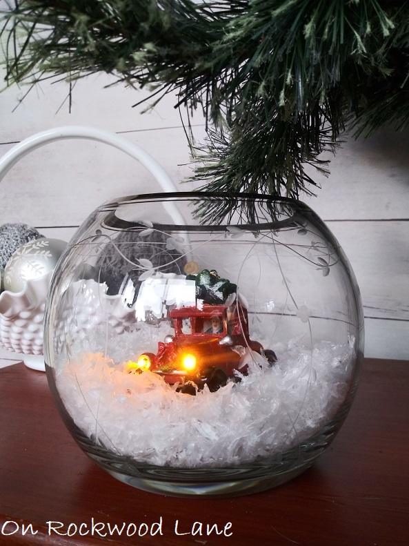 Round glass jar filled with fake snow and a small red pickup truck with lit headlights, set on a galvanized stand