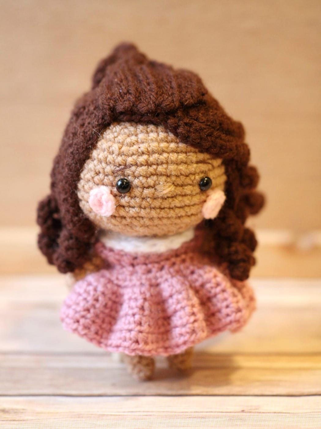 A crocheted mini doll with brown curly yarn hair, pink felt cheeks, black safety eyes and a ruffled pink dress on wood