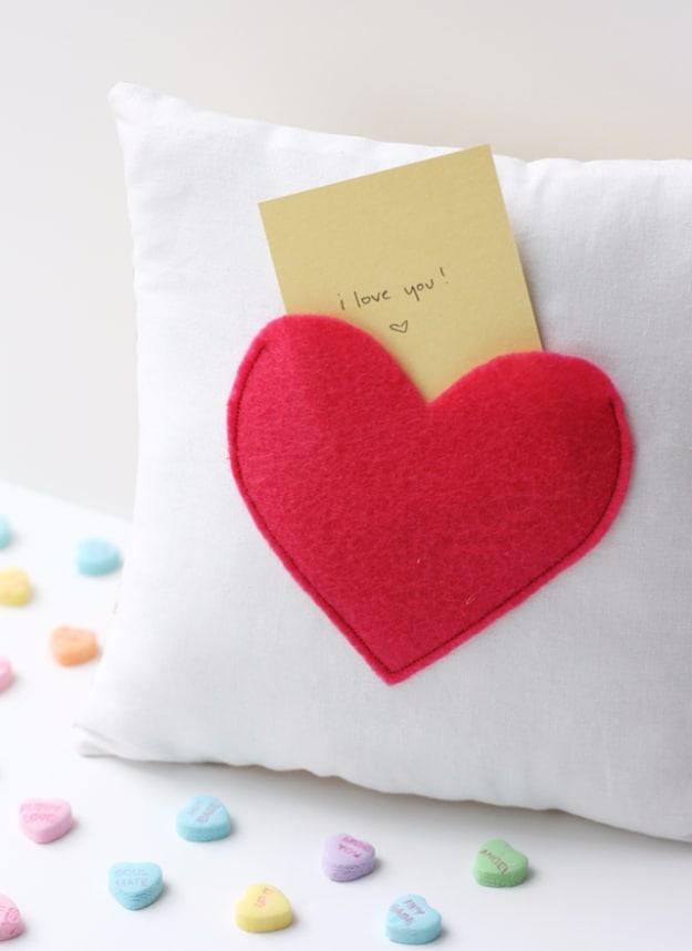White pillow with a red felt heart stitched on as a pocket holding a folded note, with candy hearts scattered nearby