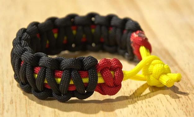 Black paracord bracelet with a red two color core and yellow cord ends, resting on a pale wooden table