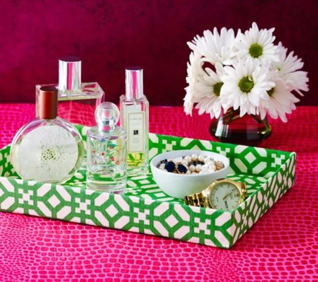 Shallow box covered in green and white geometric paper holding perfume bottles, a watch and white daisies