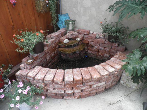 Curved low brick wall forming a corner pond and waterfall on a patio, planted with flowers around the edge