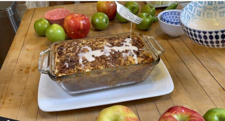 Glazed Apple Bread - white glaze being drizzled from a fork over the baked loaf in its glass dish, with sliced apples and mixing bowls behind