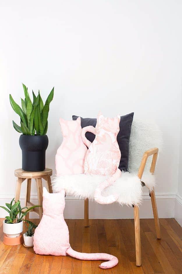DIY no sew cat pillow, three pink cat shaped pillows with long tails on a chair and floor beside a snake plant