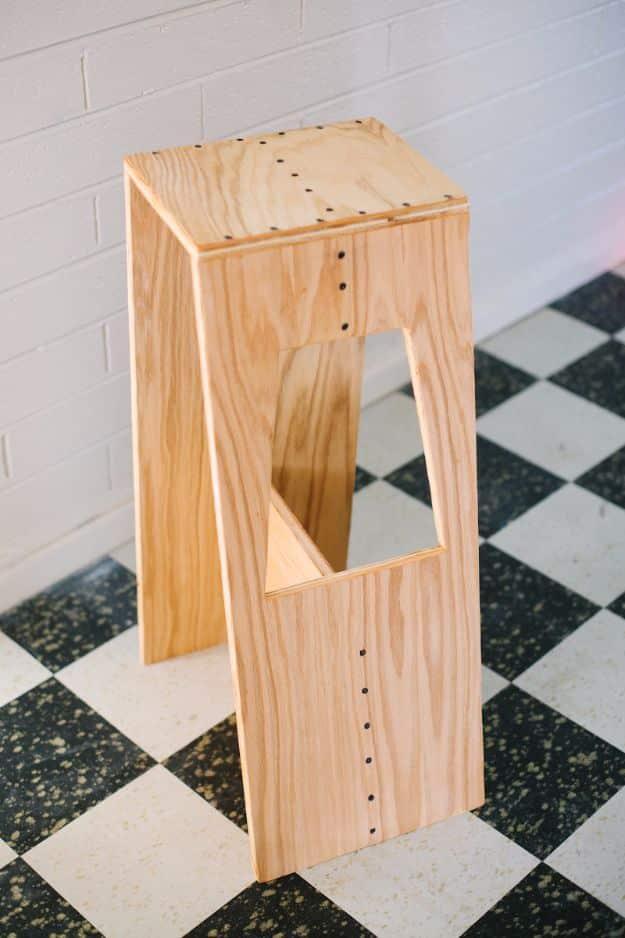 DIY modern stool made from pale plywood panels with visible screws and a cut out handle, on a checkerboard floor