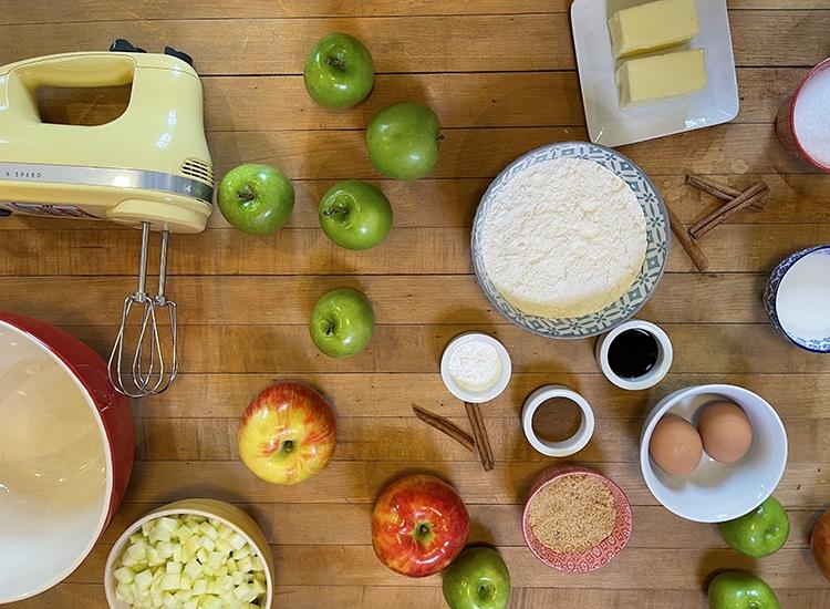 Quick Bread Recipes - flat lay of a yellow hand mixer, whole and chopped apples, flour, butter, eggs, brown sugar, cinnamon sticks and a red mixing bowl on a wood table