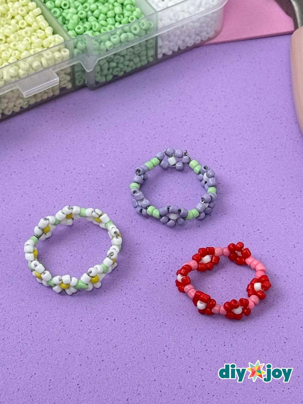 Three seed bead daisy rings in white and yellow, lavender and green and red and pink on a purple mat beside a bead box
