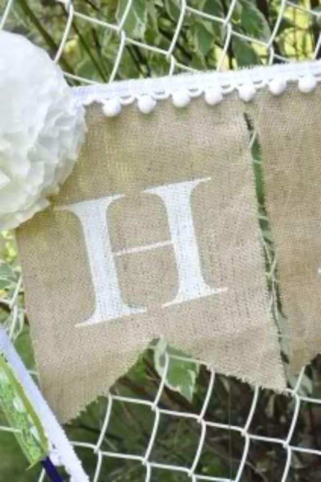 Burlap pennant with a white painted letter H and white pom pom trim, hanging against green leaves outdoors