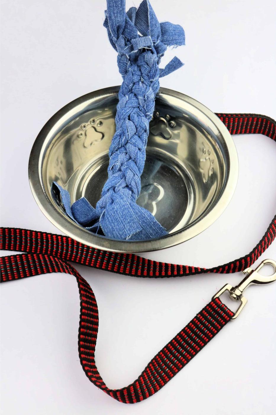 Braided blue denim dog toy standing upright in a metal dog bowl beside a red plaid leash on a white surface