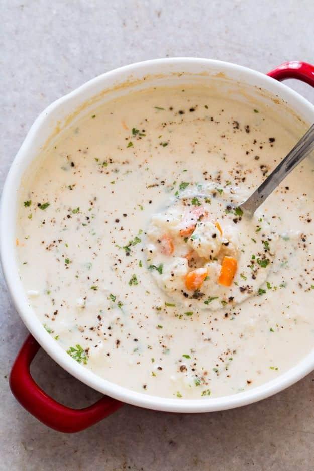 Creamy cauliflower chowder flecked with parsley and black pepper in a red handled white pot with a spoon lifting carrot