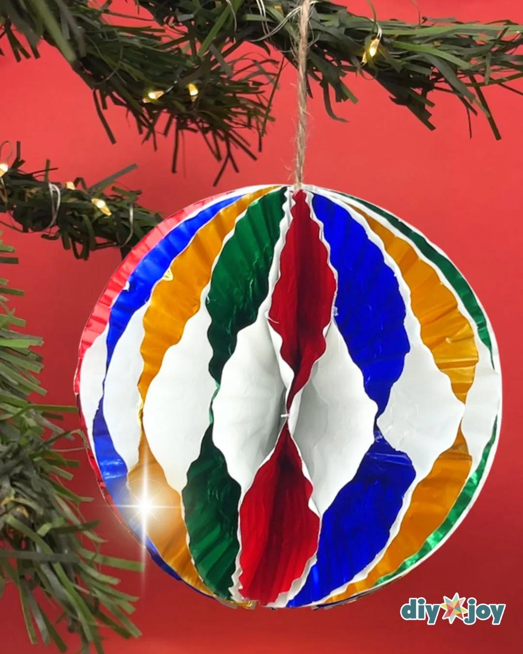 DIY Crafts - a finished striped cupcake liner ornament hanging by jute string in front of a pine garland with fairy lights