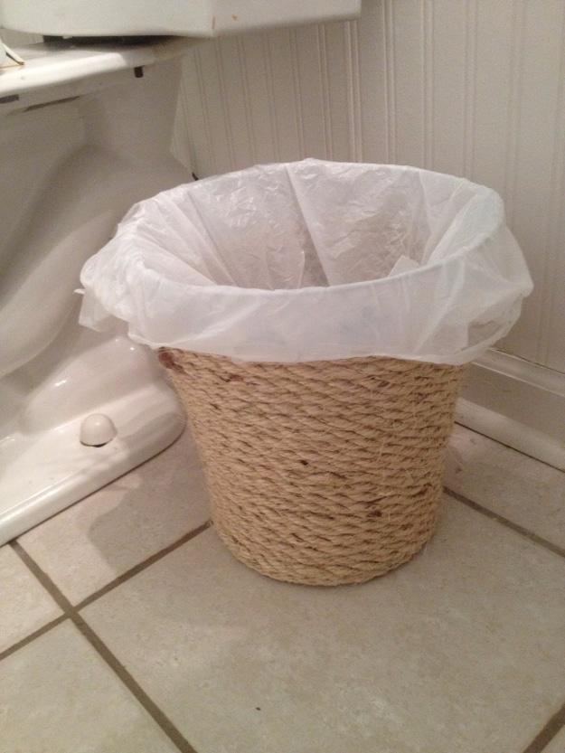 White plastic bathroom waste basket wrapped in sisal rope, lined with a white bin bag, on a tiled bathroom floor