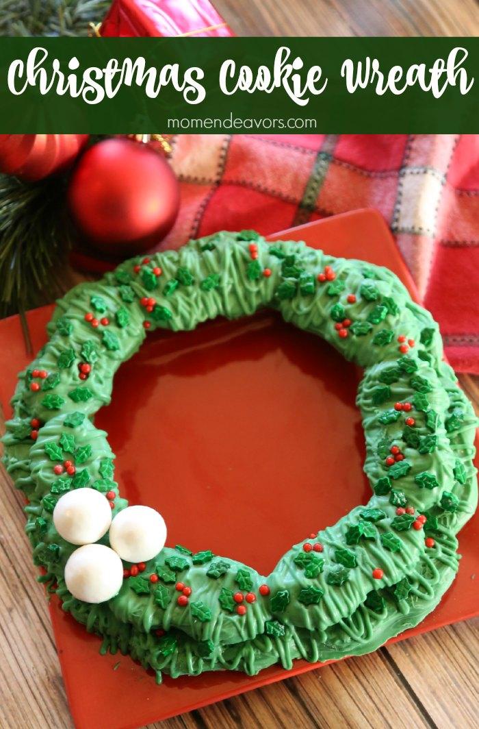 A green candy coated cookie wreath decorated with holly sprinkles, red dots and three white truffle snowballs on a red plate