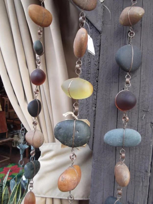 Hanging chains of smooth oval stones wrapped in copper wire, displayed against a beige curtain and a weathered post