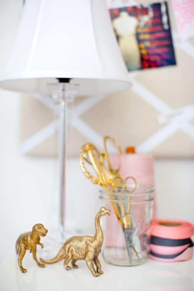 DIY gold animal figurines, two small gold sprayed plastic dinosaurs on a white desk beside a jar of gold scissors