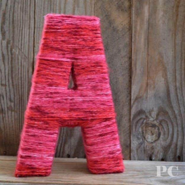DIY baby shower decoration yarn wrapped letter A in variegated red and pink yarn standing against weathered grey boards