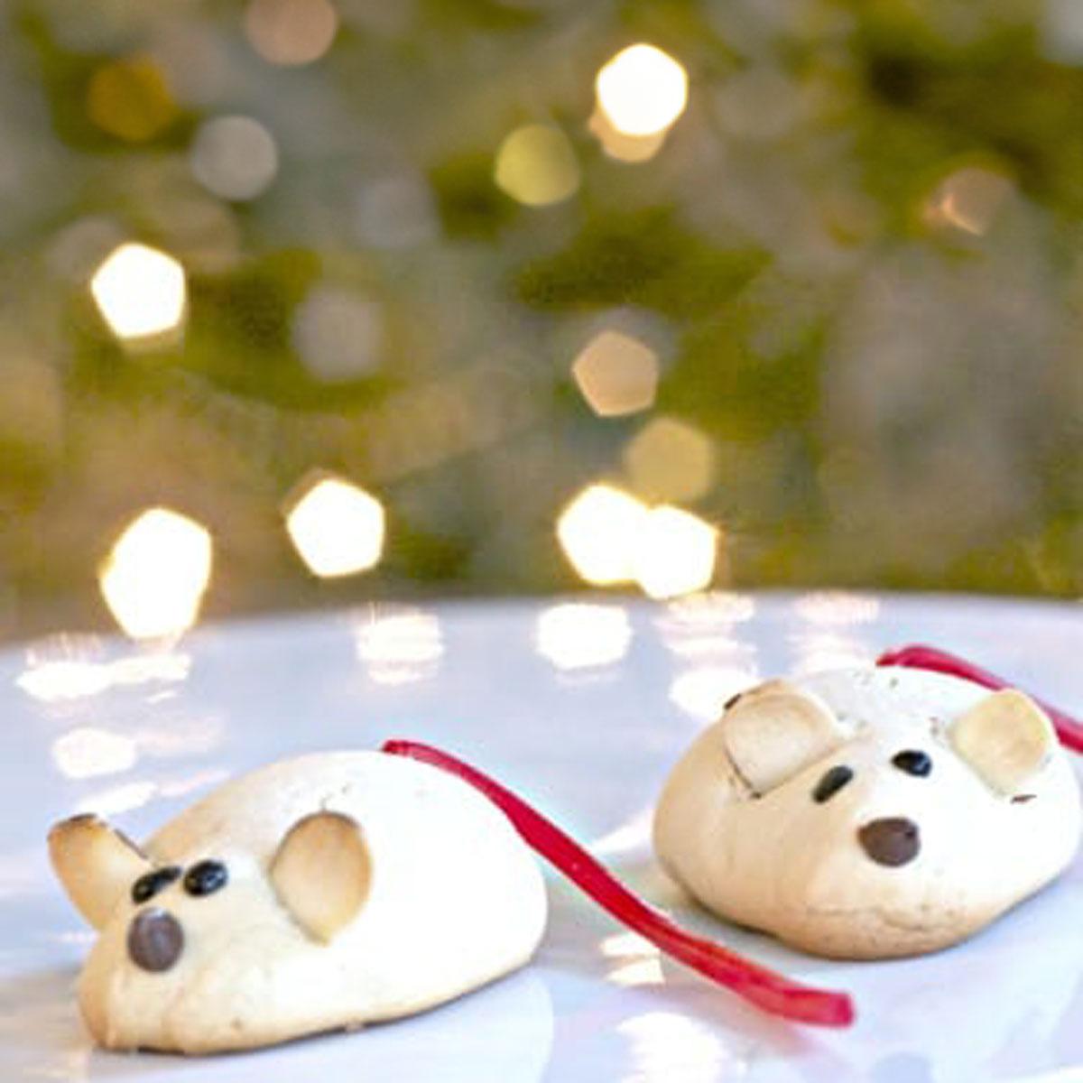 Two mouse shaped almond cookies with sliced almond ears, chocolate chip noses and red licorice tails on a plate by a lit tree