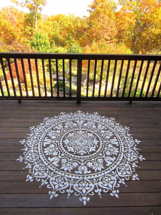 White mandala pattern stenciled onto brown deck boards outdoors with autumn trees beyond the railing