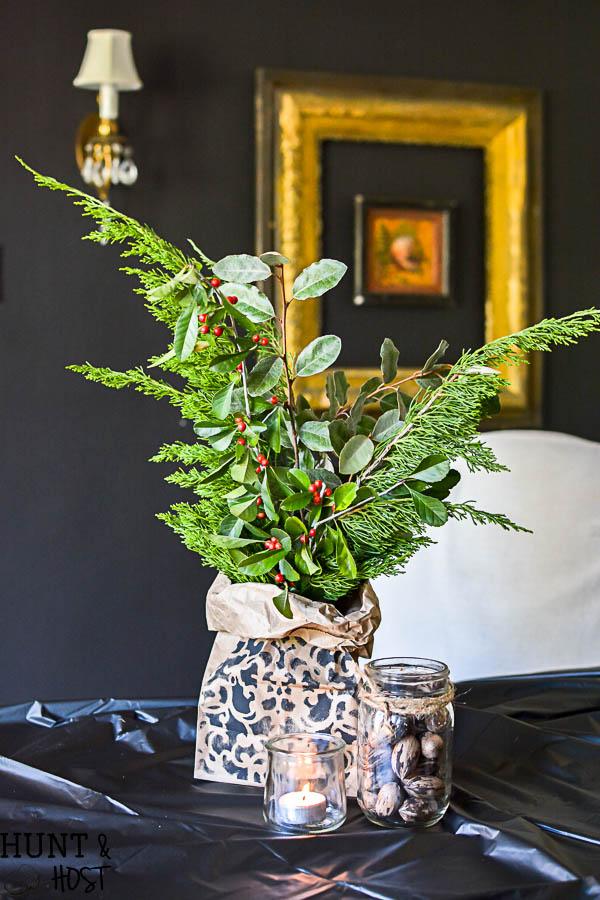 A stenciled brown paper sack vase full of juniper, silver leaves and red berries beside a votive candle and a jar of nuts