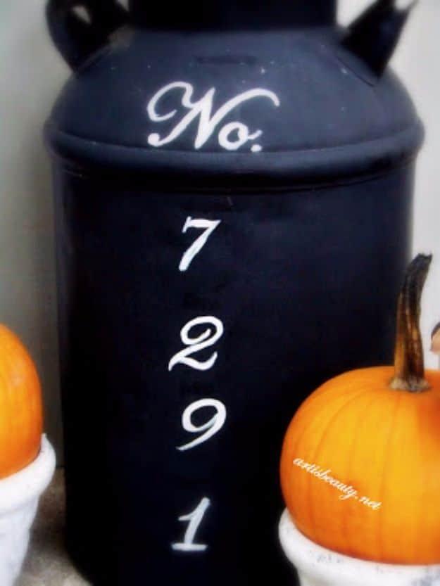 Black painted antique milk can with white hand lettered house numbers, standing on a porch with pumpkins