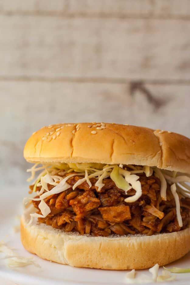 Pulled BBQ jackfruit sandwich piled with coleslaw on a sesame seed bun against a rustic wood background