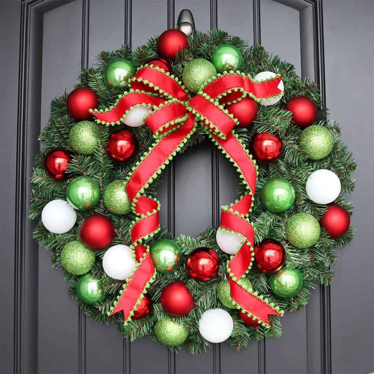 An evergreen wreath covered in red, green and white ball ornaments with a red bow trimmed in green pom poms on a dark door