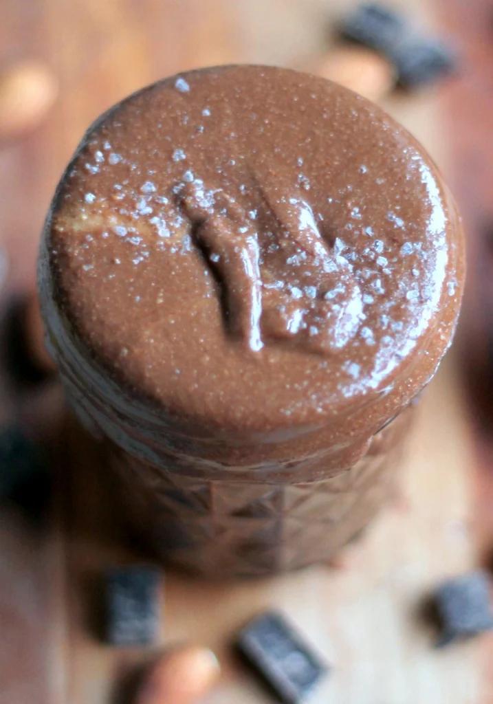 Glass jar of glossy dark chocolate almond butter with a spoon lifting some out and almonds scattered nearby