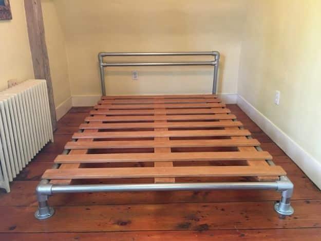 Pipe and flange bed frame with a galvanized pipe headboard and wood slats in a sunlit wood floored room