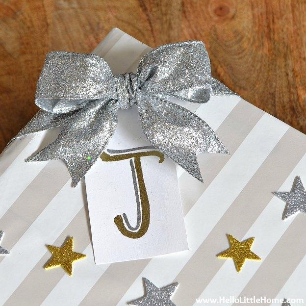 A striped wrapped gift with a big silver glitter bow, a gold monogram tag and scattered glitter paper stars