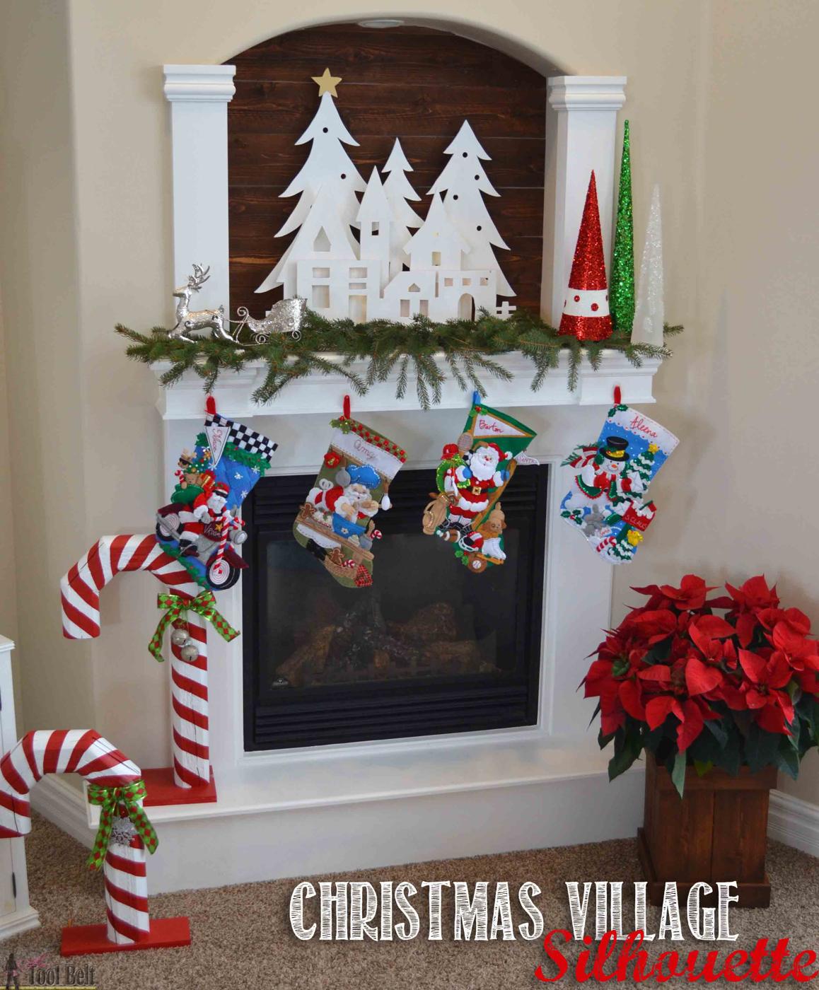 White layered plywood Christmas village silhouette with trees and houses on a mantel with stockings and a poinsettia