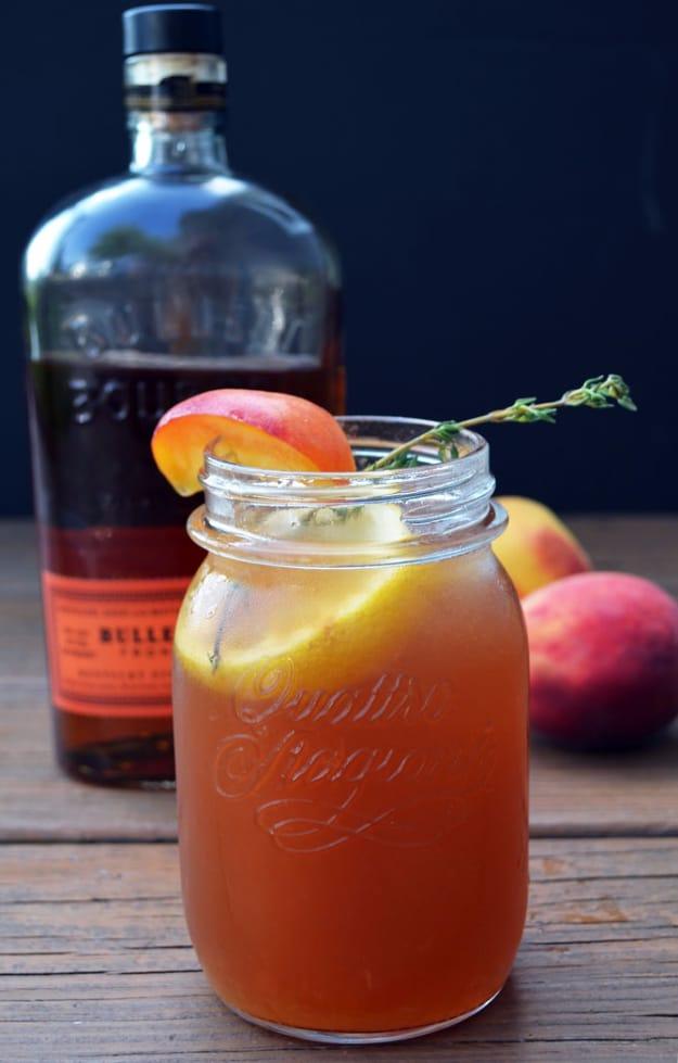 A mason jar of bourbon peach sweet tea garnished with thyme beside a bourbon bottle and fresh peaches
