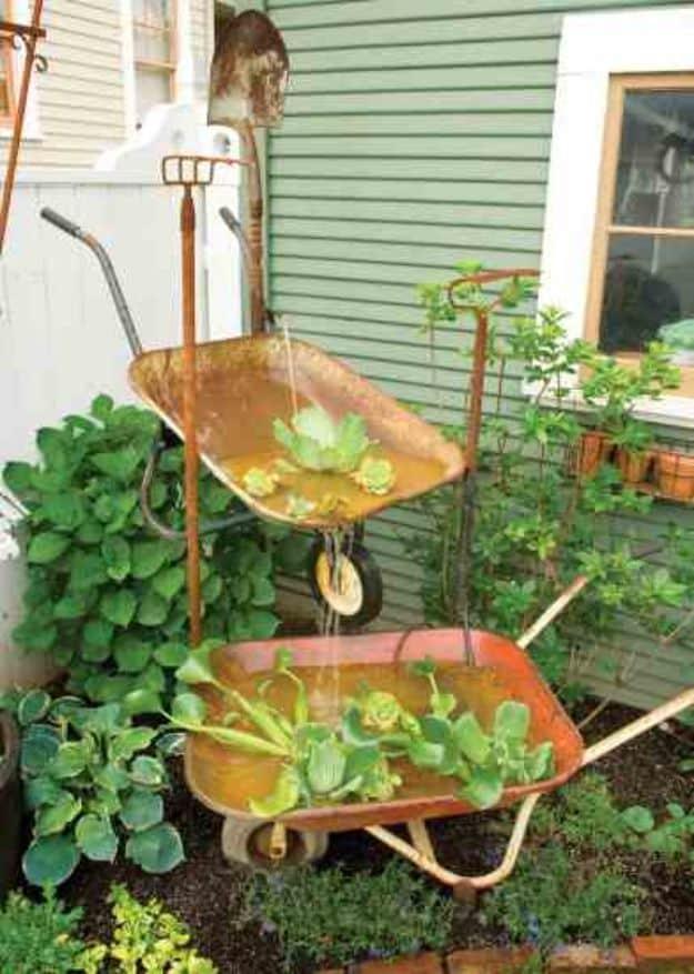 Two rusted wheelbarrow fountains planted with water plants beside a green house wall, one on a stand