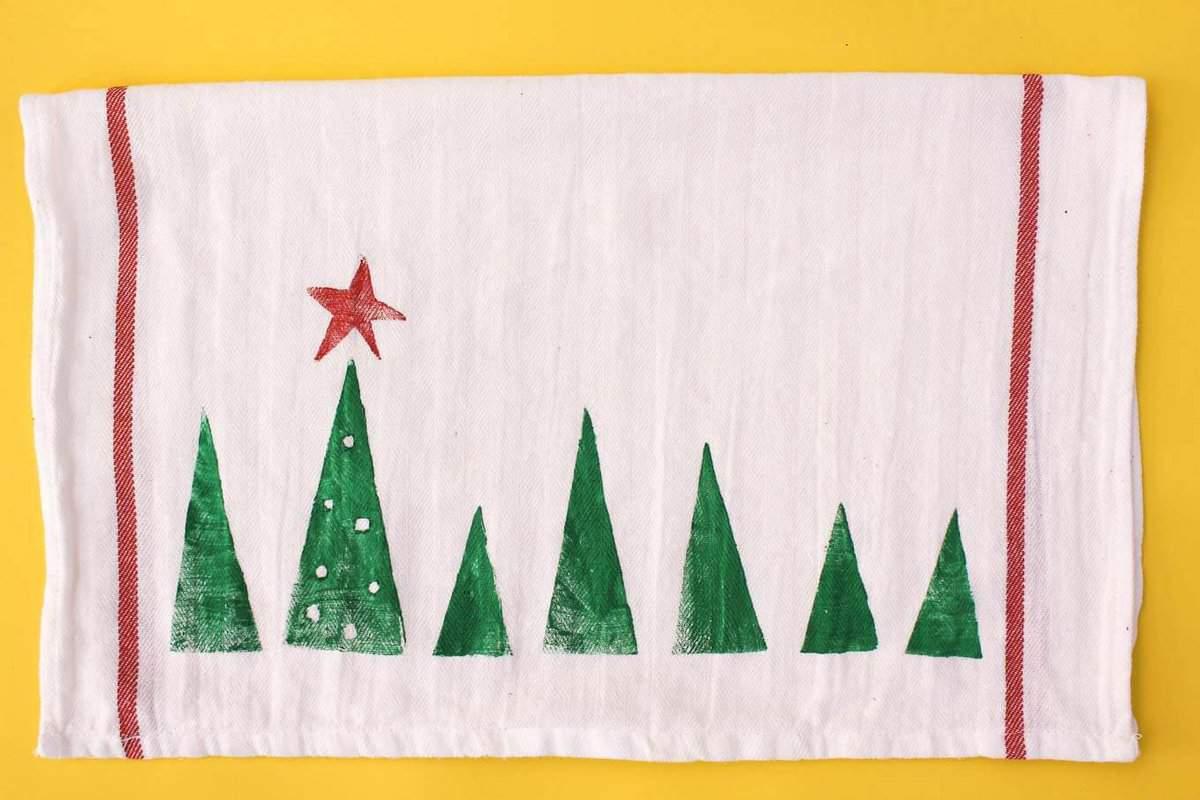 White flour sack tea towel stamped with a row of green potato print Christmas trees and a red star on a yellow background