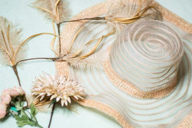 Wide brimmed straw hat trimmed with cream ribbon, a peach flower, feathers and small blooms on a pale surface