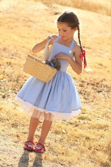 Child wearing a blue gingham Dorothy dress with red shoes and woven basket outdoors with Girl Inspired source mark