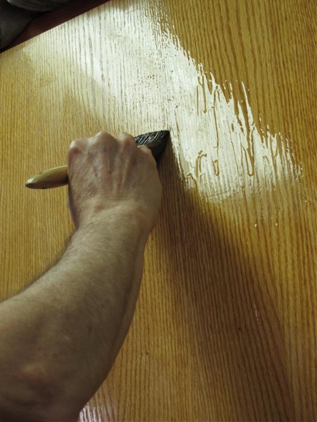 Brush working an amber finish across a wood door panel, the wet surface catching light across the grain