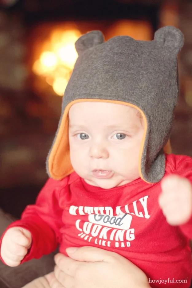Baby in a grey fleece hat with round bear ears and orange lined ear flaps, in front of a lit fireplace