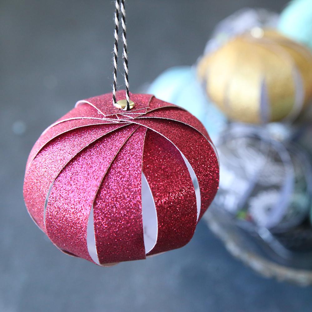 A magenta glitter paper strip ball ornament hanging on black and white twine with gold and aqua paper balls blurred behind it