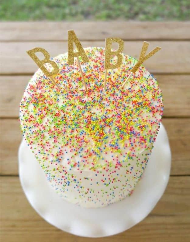 White baby shower cake completely covered in rainbow nonpareil sprinkles with a gold glitter BABY cake topper