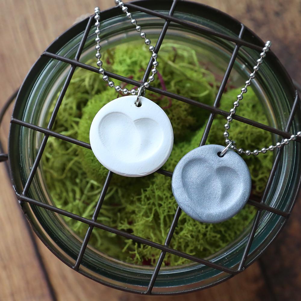 Two round clay fingerprint heart pendants, one white and one silver, on ball chains against green moss