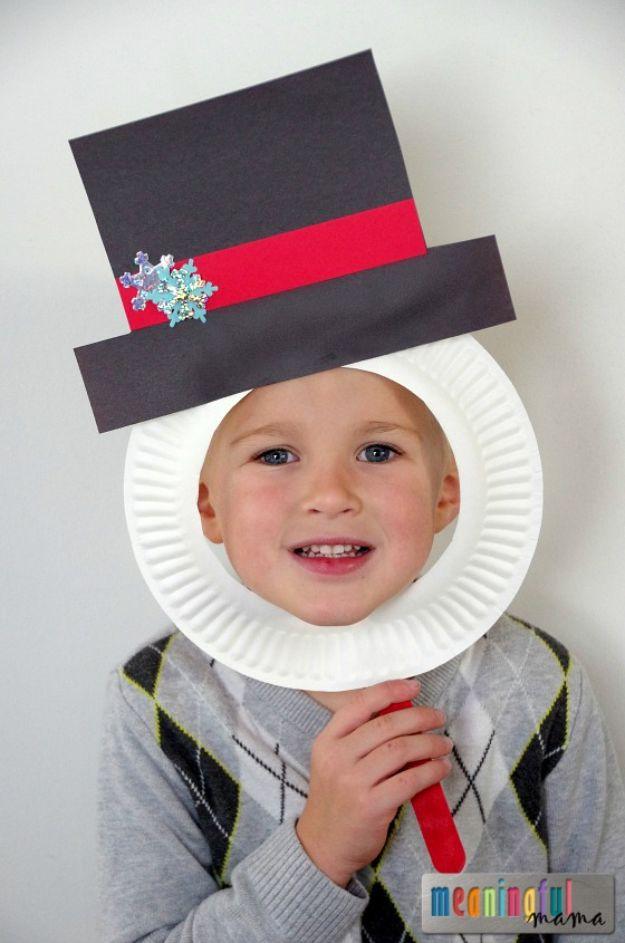 A boy holding a paper plate mask to his face with a black paper top hat, red band and a snowflake sticker