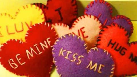 A pile of stuffed felt hearts in red, purple, yellow and cream with blanket stitched edges, reading be mine and kiss me
