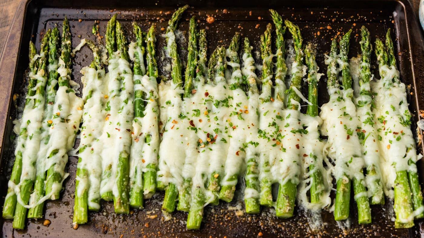 Cheesy asparagus spears on a sheet pan under melted mozzarella and parmesan, with red pepper flakes on top