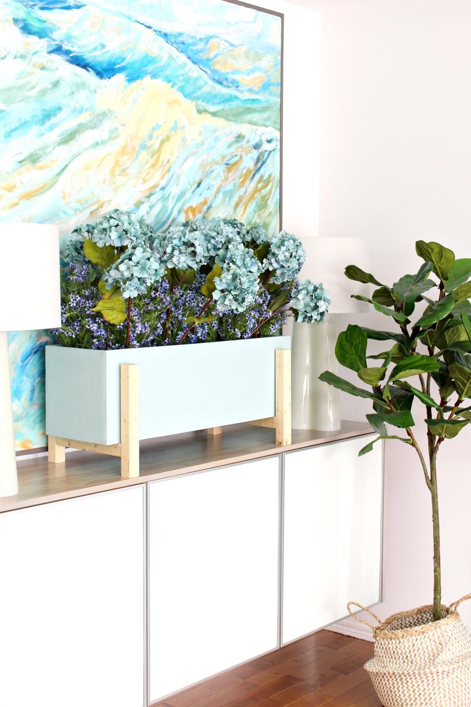 Modern plywood planter box, a pale blue rectangular planter on light wood legs filled with blue hydrangeas on a white console