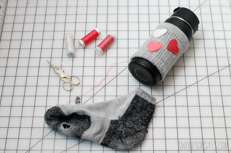 Gray ribbed sock cuff coffee cozy with three red felt hearts on a black travel cup, beside scissors, floss and the cut sock